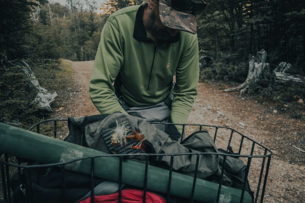 A man in a green jacket organizes outdoor gear, including fishing items, in a basket on a forested trail.
