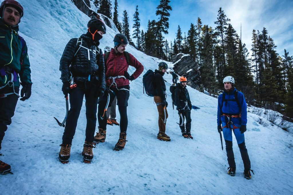 A group of six climbers, equipped with ice axes and crampons, stands on a snowy slope surrounded by tall evergreen trees.