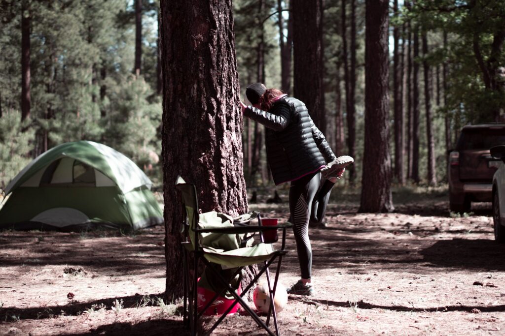 A woman stretches against a tree in a forest campsite, with a green tent and camping chair nearby.