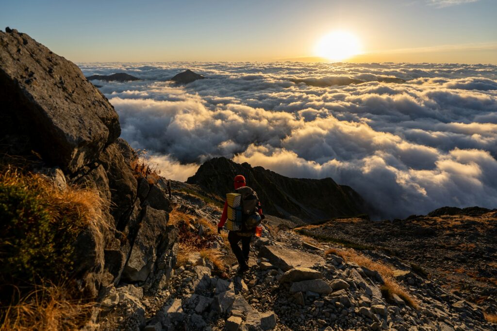 A hiker descends rocky terrain above clouds, bathed in golden sunlight at sunrise, with mountains silhouetted in the background.