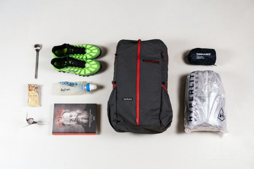 A flat lay of hiking gear including green athletic shoes, a black backpack, a water bottle, snacks, a book, and a sleeping bag.