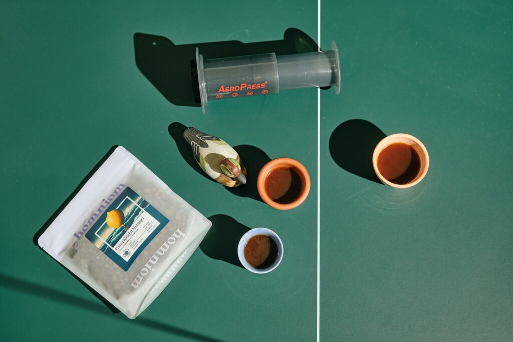 An AeroPress, two coffee cups, a bag of coffee beans, and a decorative bird figurine on a green table.