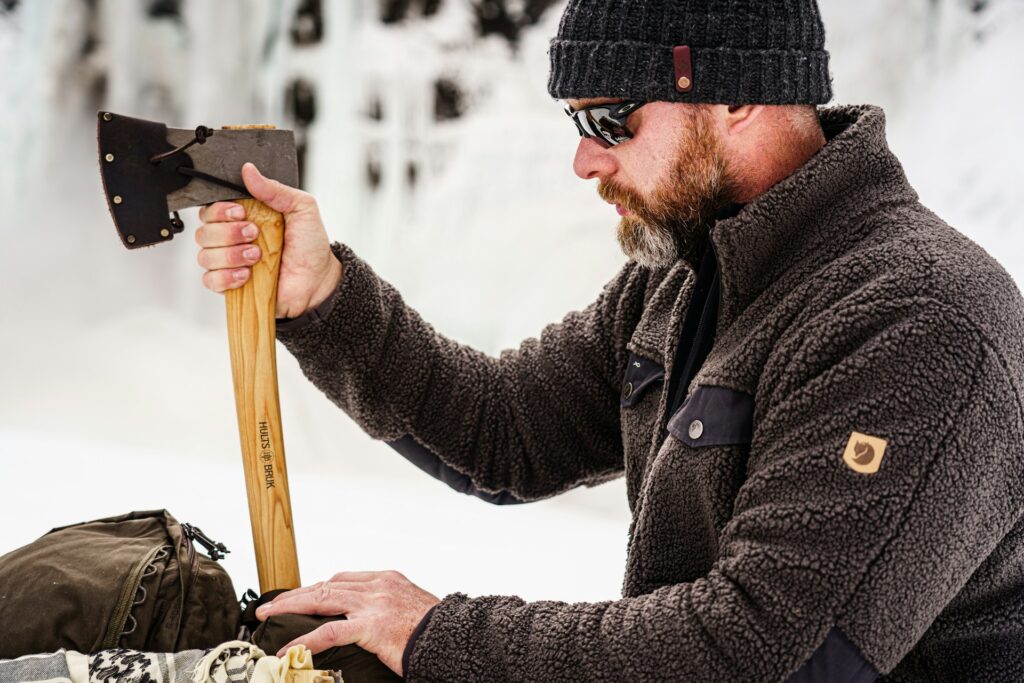 A person in a brown fleece jacket and knit hat holds an axe above a neatly arranged outdoor gear against a snowy backdrop.