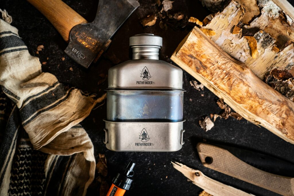 A stainless steel Pathfinder water bottle surrounded by an axe, knives, wood, and fabric, suggesting an outdoor adventure theme.