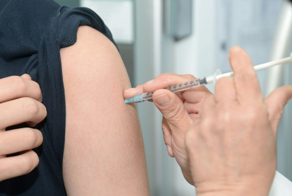 A healthcare professional administers a vaccine to a patient's arm using a syringe. The arm is partially exposed and relaxed.