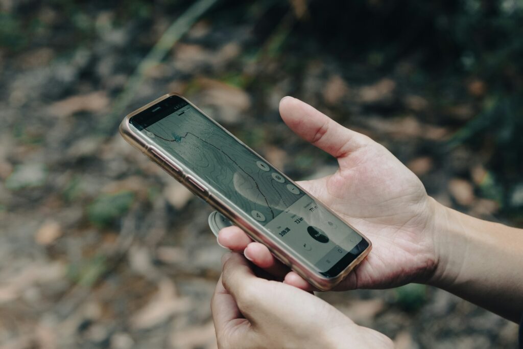 A person holds a smartphone displaying a topographic map, surrounded by nature with leaves in the background.