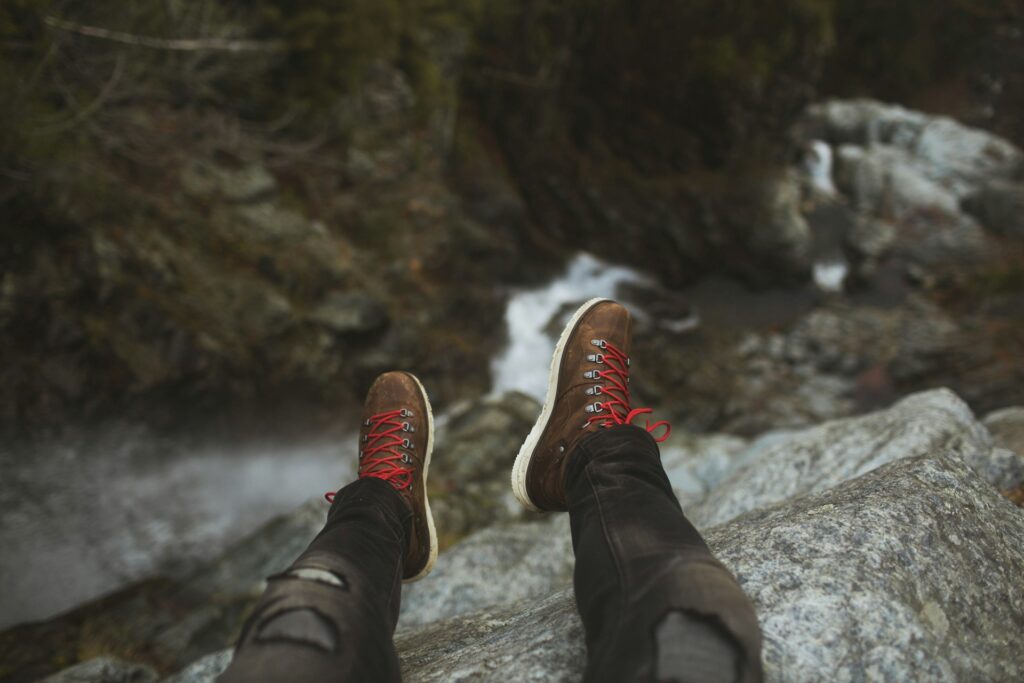Person's legs in torn jeans and hiking boots with red laces dangling over the edge of a rocky cliff, above a flowing stream below.