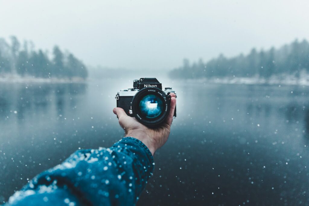 A person holds a Nikon camera in front of a snowy river, surrounded by a misty landscape of trees and falling snowflakes.