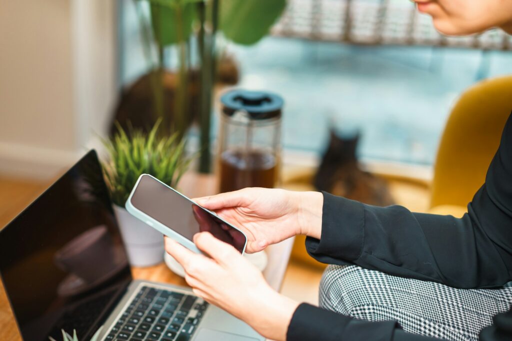 A person in a black shirt holds a smartphone, sitting beside a laptop, plants, and a coffee maker, with a cat in the background.