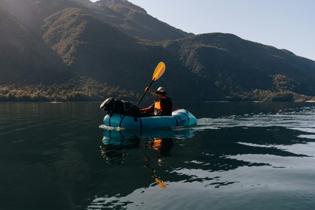 A person paddles a bright blue kayak on a calm lake, surrounded by lush mountains under a clear sky.