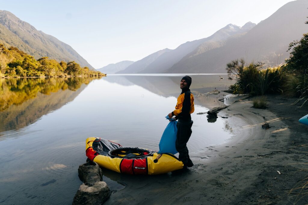 A person stands by a calm lake in the morning, holding a blue bag next to a colorful kayak, with mountains reflected in the water.