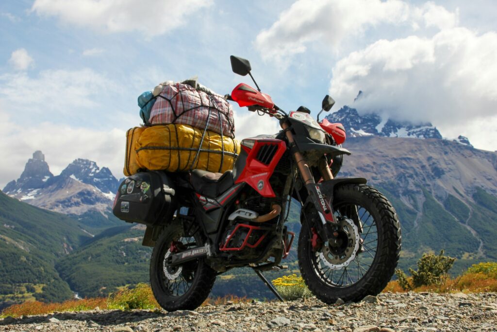 A red motorcycle loaded with colorful bags stands against a backdrop of majestic mountains and a blue sky, ready for adventure.