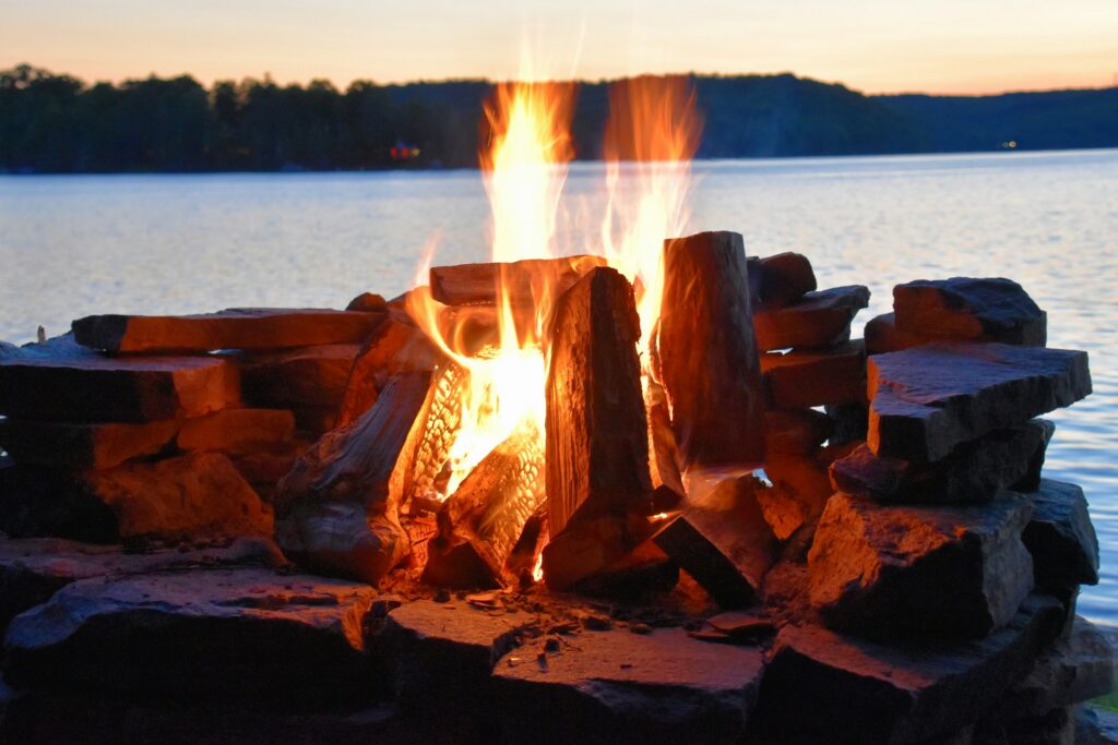 A glowing campfire crackles among stacked stones, with a serene lake and twilight sky in the background.