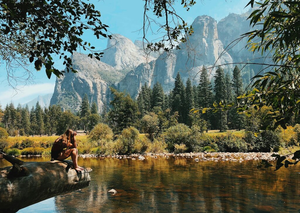 A woman sits on a log by a serene river, surrounded by lush trees and majestic mountains under a clear blue sky.
