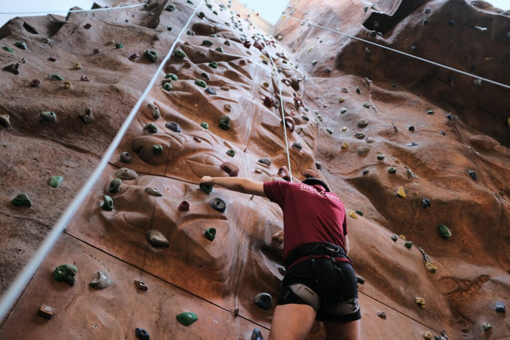 A climber in a red shirt ascends a textured indoor rock climbing wall, gripping colorful holds while secured by a safety rope.
