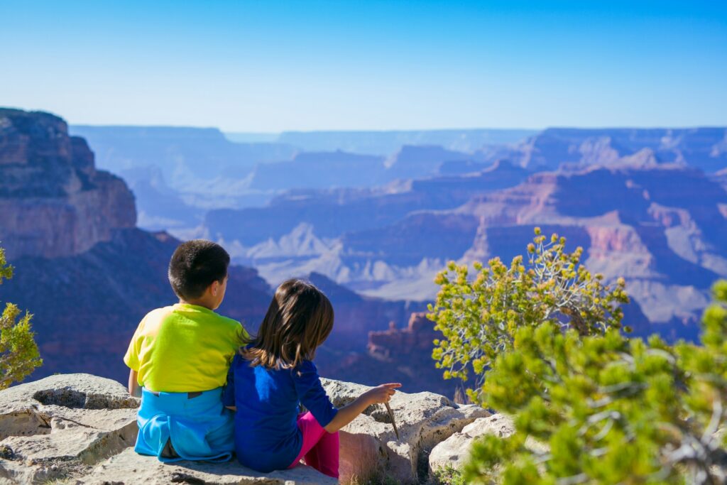 Two children sit on a rocky ledge, gazing at the vast Grand Canyon landscape under a clear sky.