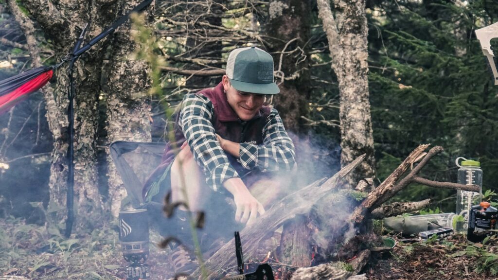 A person sits on the ground near a campfire surrounded by trees, smoke rising, with camping gear scattered nearby.
