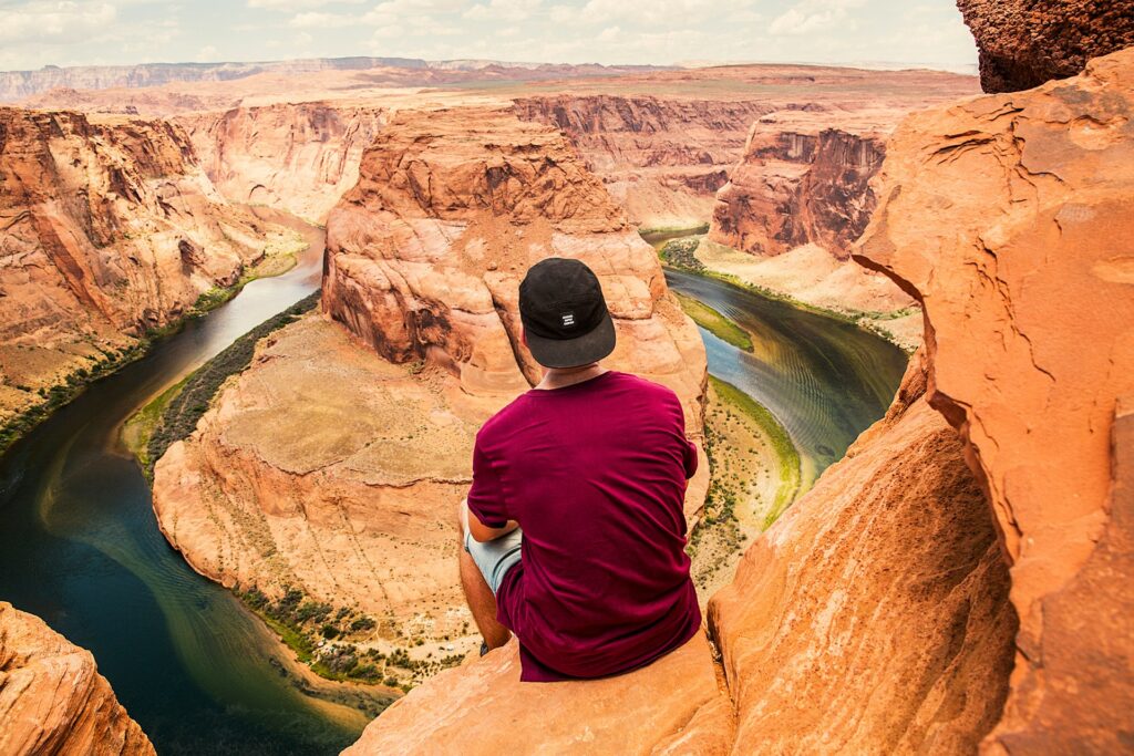 A person sits on a rock ledge overlooking the Horseshoe Bend, with winding blue-green water surrounded by red rock formations.