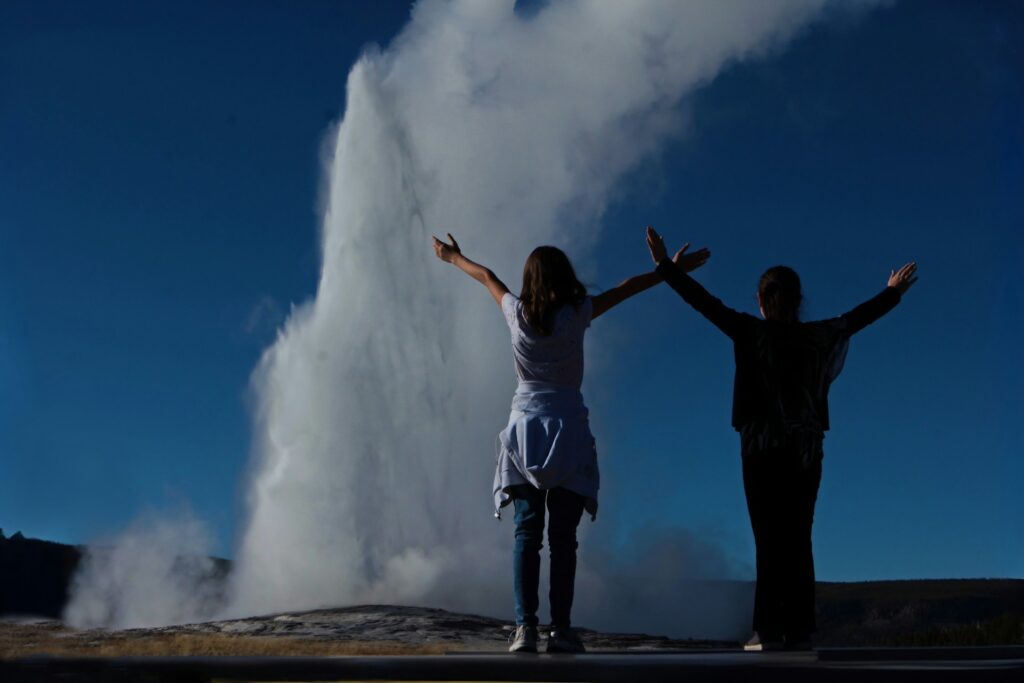 Two women raise their arms in joy as Old Faithful geyser erupts in the background, against a clear blue sky.