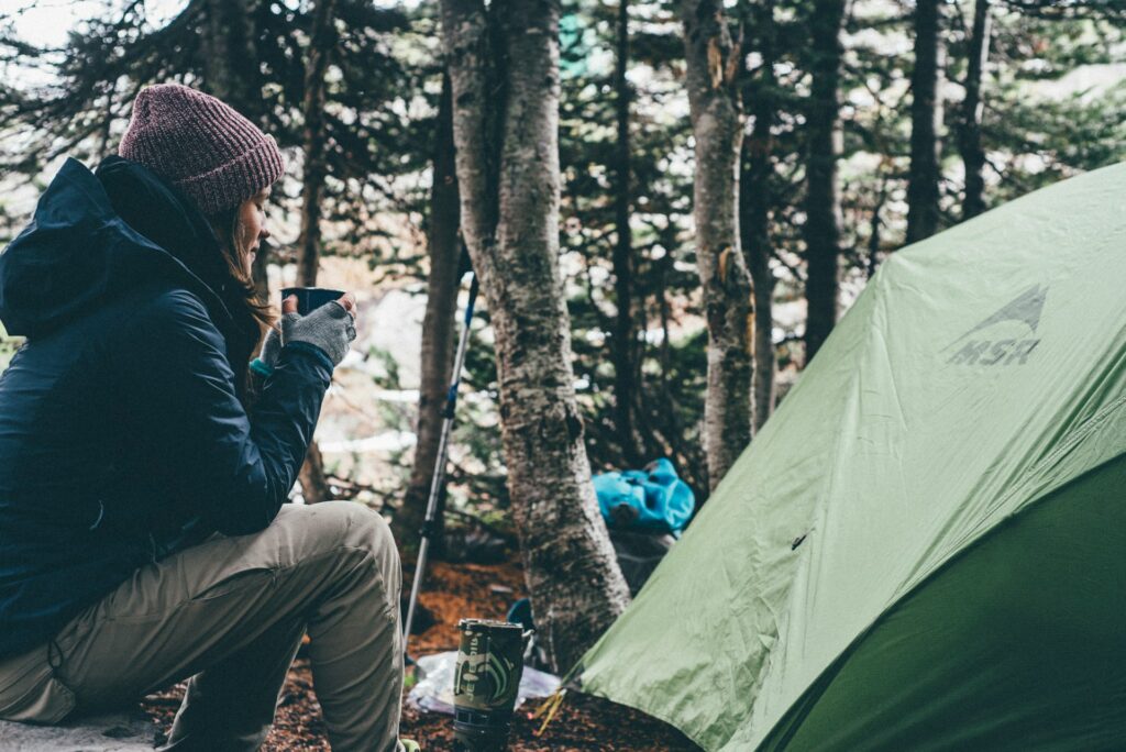 A person sits beside a green tent, enjoying a drink surrounded by trees in a peaceful camping setting.