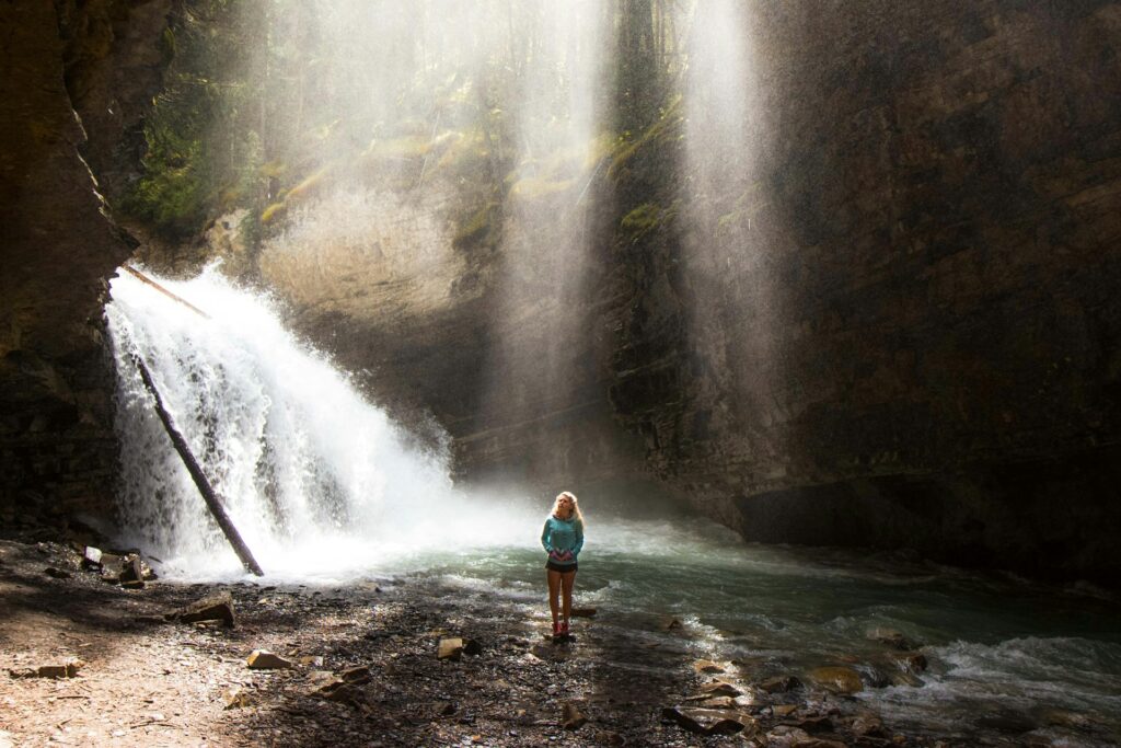 A person stands near a powerful waterfall, mist rising in a sunlit, rocky gorge surrounded by lush greenery.