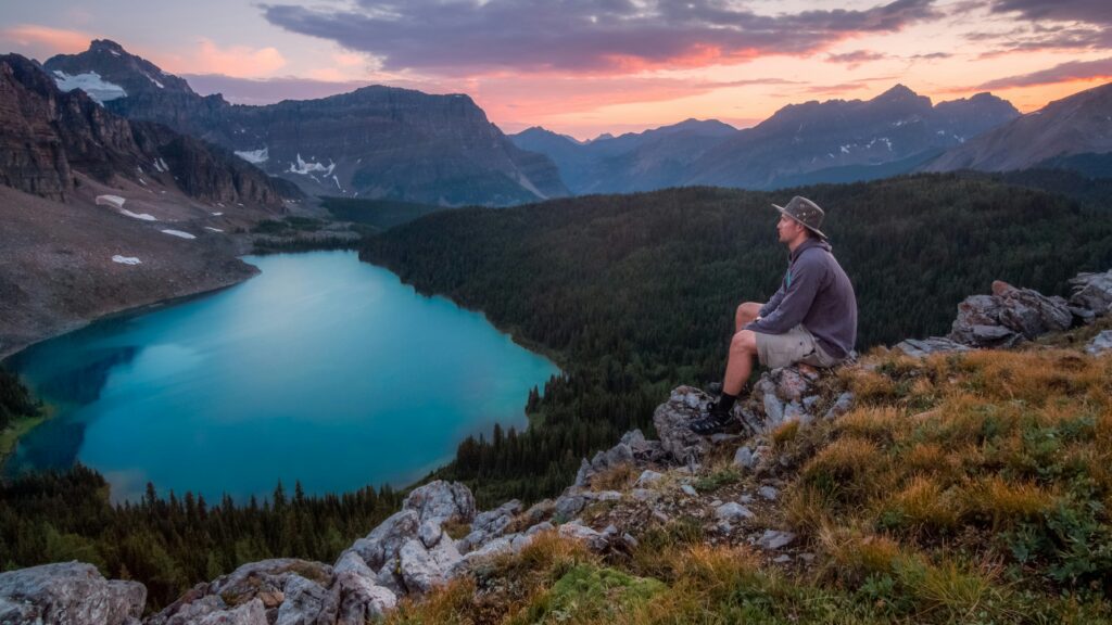 A person sits on a rocky ledge overlooking a tranquil turquoise lake, framed by lush forests and majestic mountain peaks at sunset.