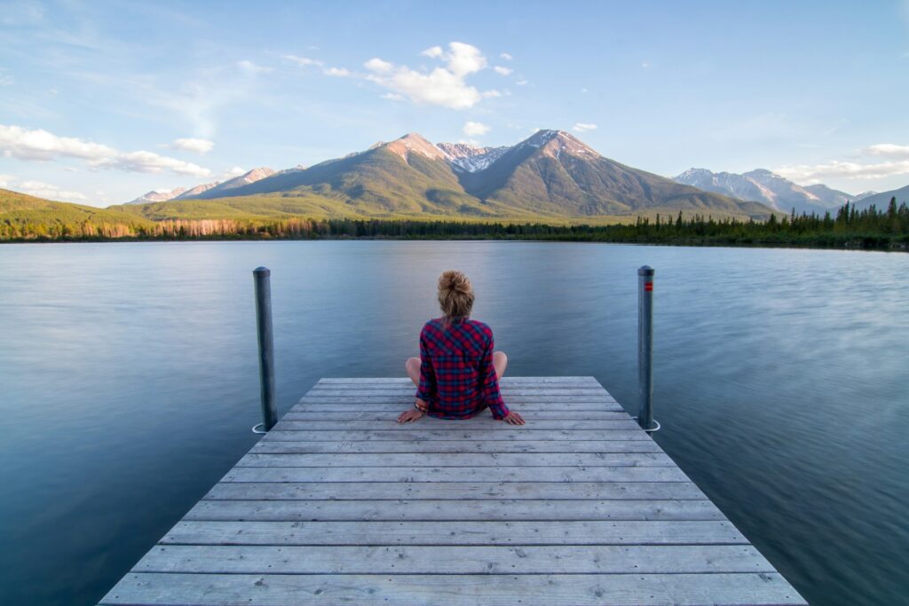 A person sits on a wooden dock, gazing at serene mountains and a calm lake under a clear sky.
