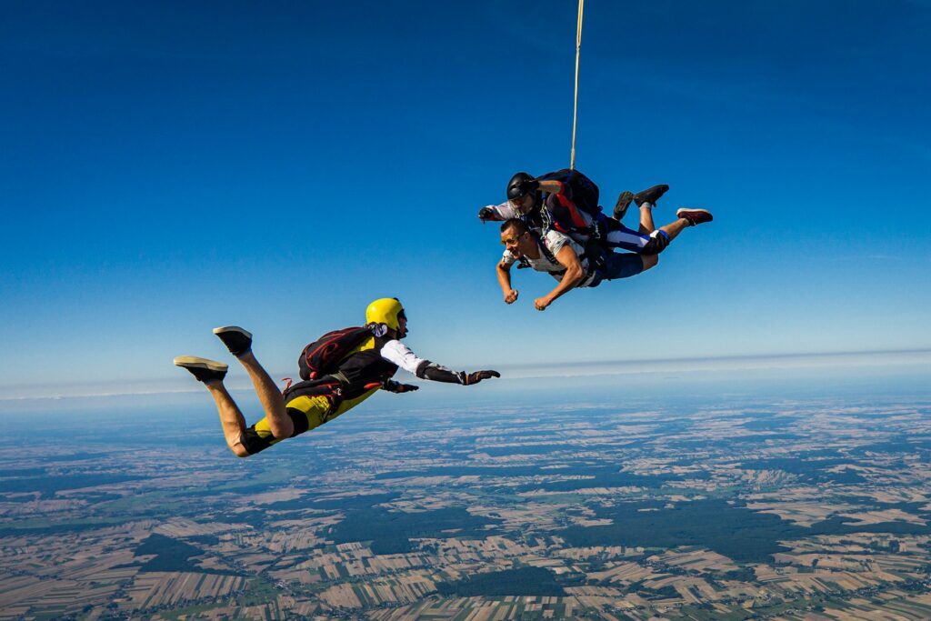 A tandem skydive in action, with parachutists soaring above a patchwork of green and brown fields against a bright blue sky.