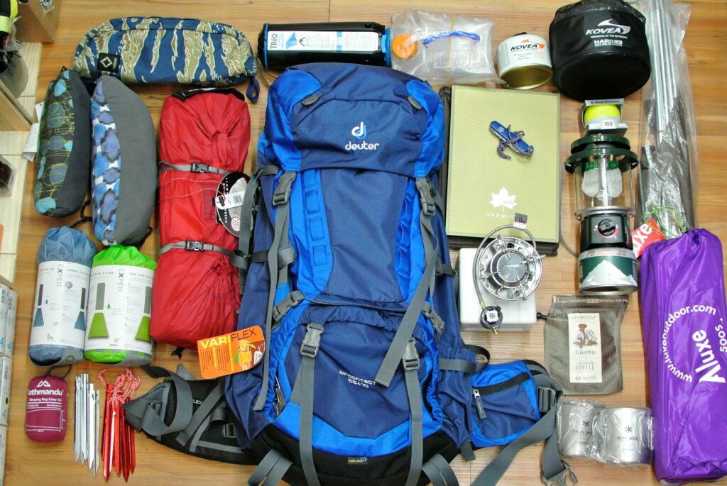A neatly arranged collection of camping gear, including a blue backpack, tents, a stove, mugs, and various camping accessories.