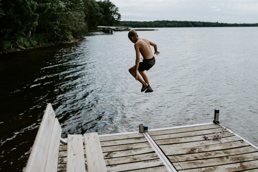 A young boy leaps from a wooden dock into calm water, surrounded by trees and a cloudy sky. Excitement and joy radiate from the moment.
