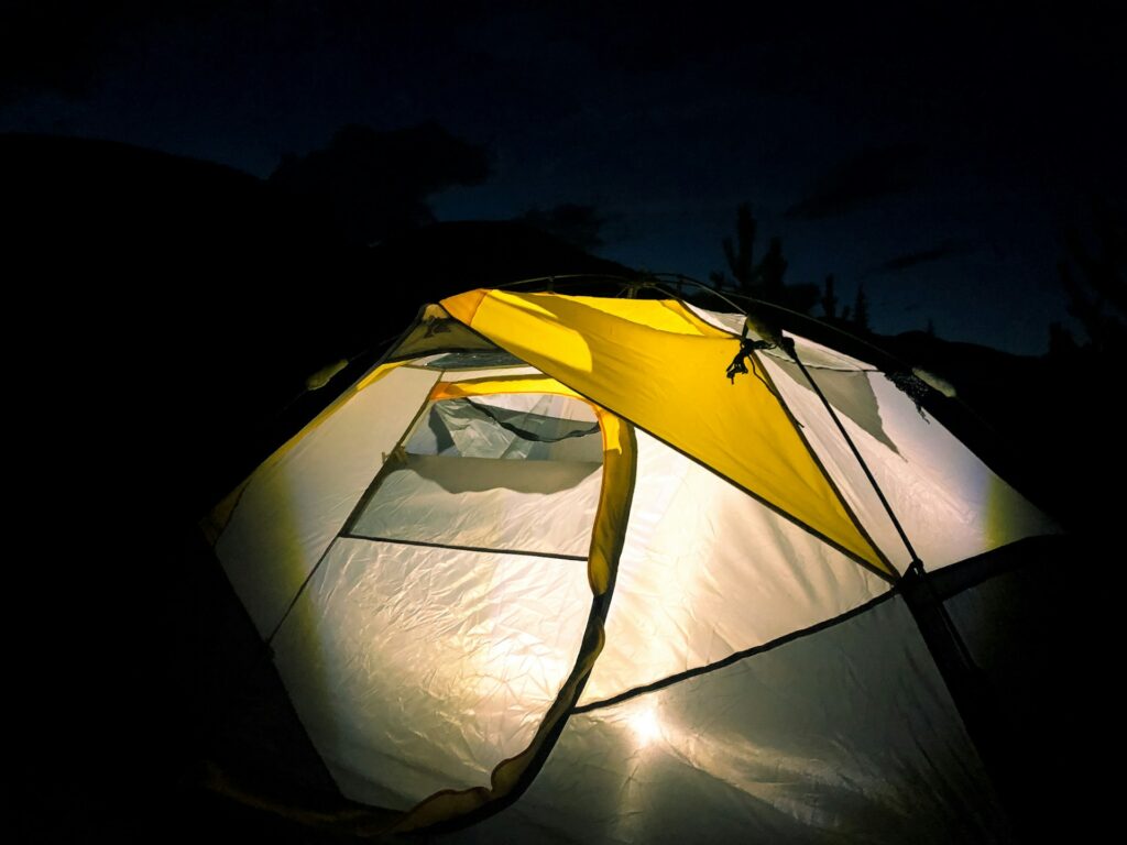 Illuminated tent in a dark campsite, with a soft yellow light shining through its fabric, surrounded by a night sky.