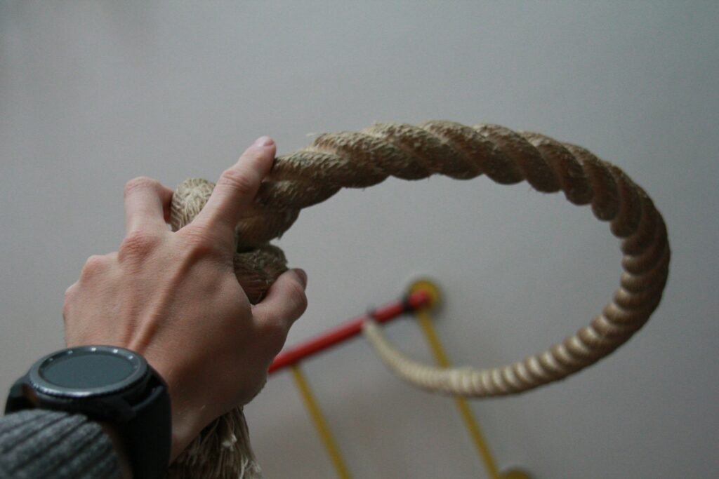 A hand grips a thick, coiled rope with a watch on the wrist; a colorful structure partially visible in the background.
