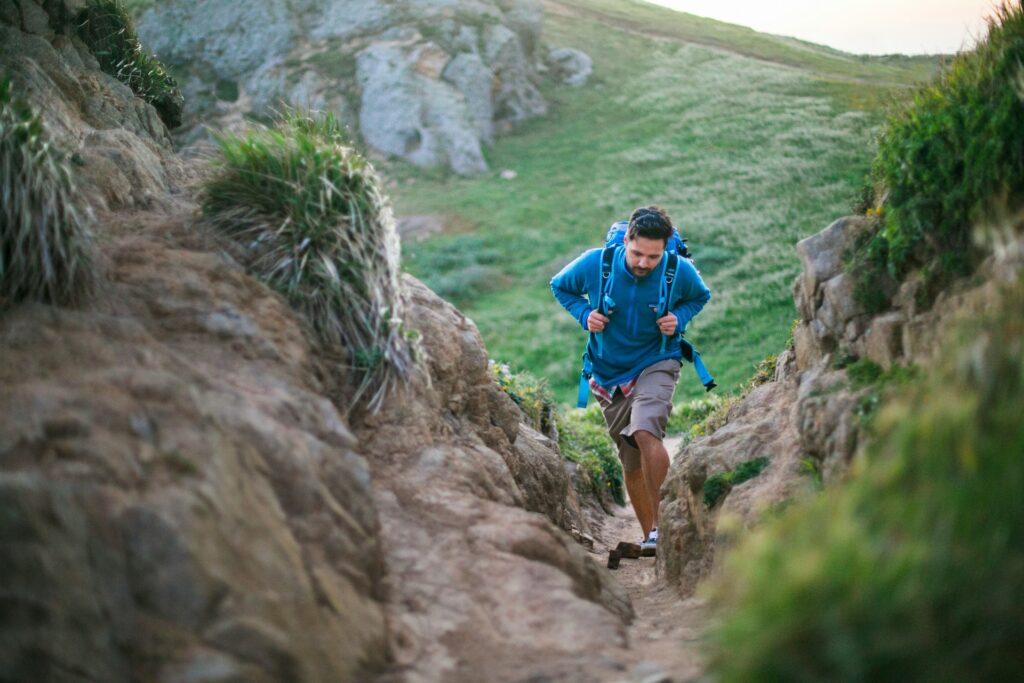 A hiker ascends a narrow rocky path surrounded by green grass and rugged terrain under a soft, natural light.
