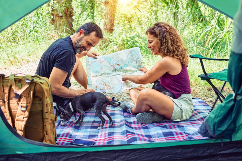 A couple sits inside a tent, examining a map on a plaid blanket, with a small dog exploring nearby, surrounded by lush greenery.