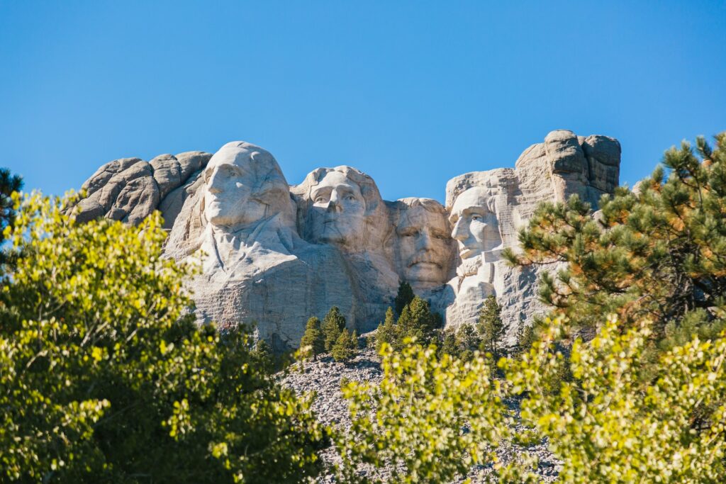 Mount Rushmore with large granite presidential sculptures set against a clear blue sky, framed by green trees in the foreground.