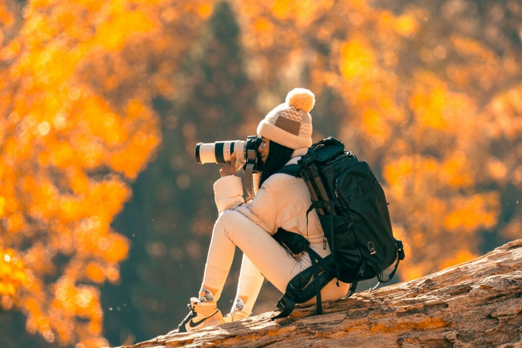 A person sits on a log, holding a camera, surrounded by vibrant autumn foliage in warm golden hues. A backpack rests beside them.
