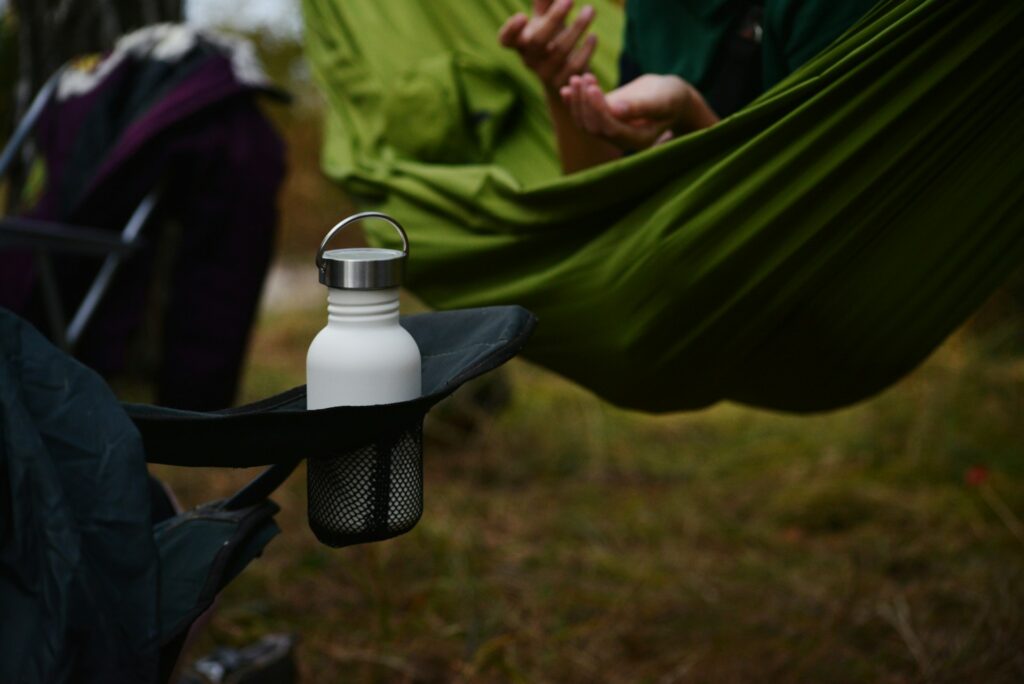 A white water bottle rests in a black mesh cup holder, with green hammocks and hands gesturing in the background at a campsite.