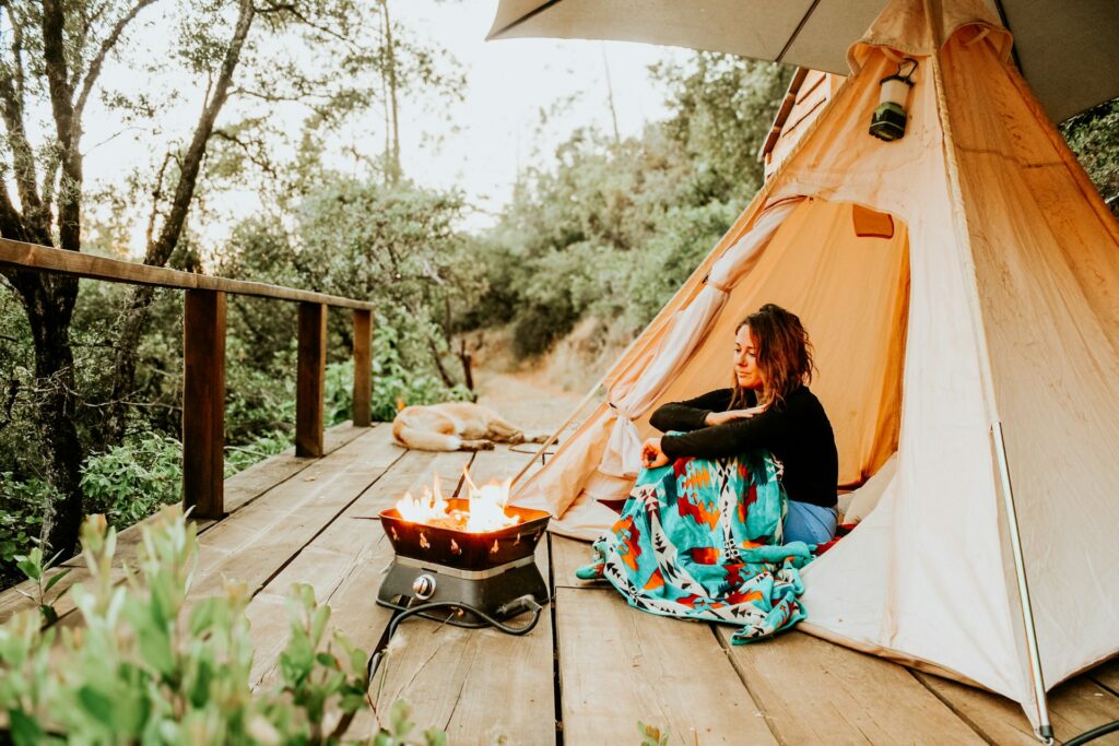 A person sits on a wooden deck by a cozy campfire outside a tent, surrounded by lush greenery and a serene natural setting.