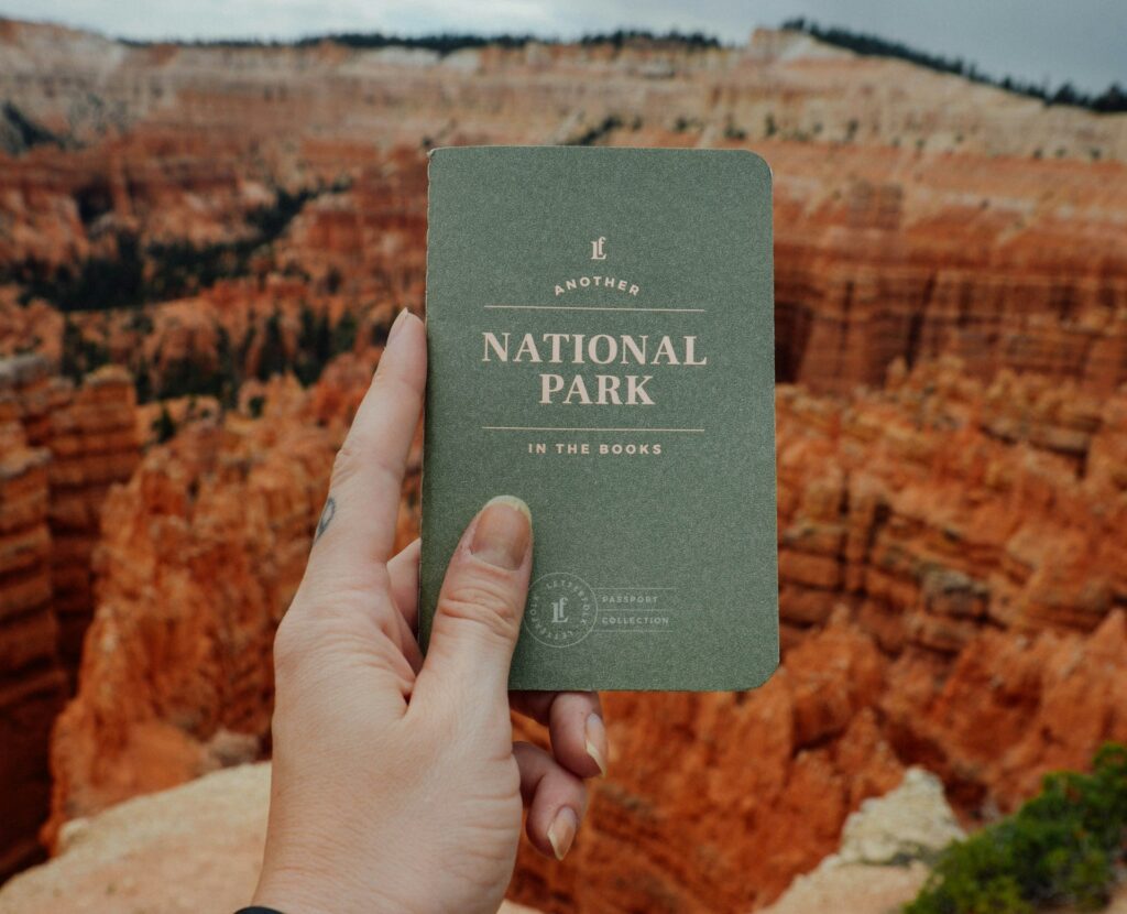 A hand holds a green booklet titled "Another National Park" against a stunning canyon backdrop. The scene captures nature's grandeur.