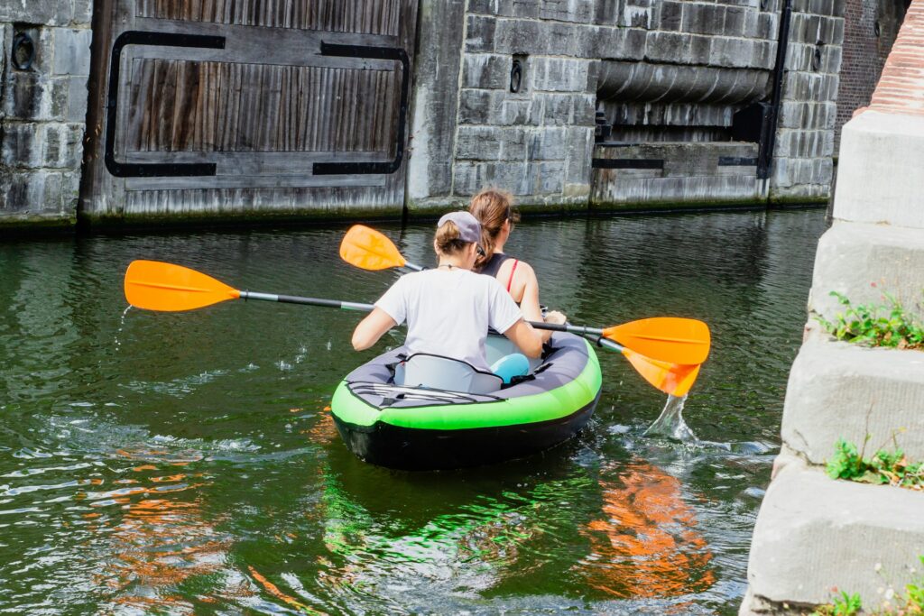Two women paddle a kayak with orange paddles in a serene canal, approaching a large wooden lock gate.