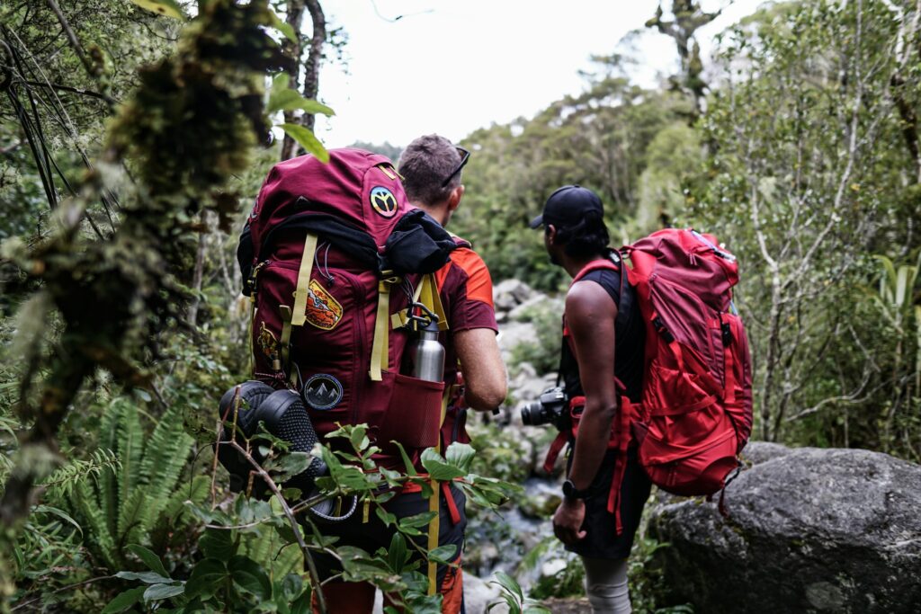 Two hikers with backpacks stand by a river in a lush forest, observing their surroundings while preparing for their adventure.