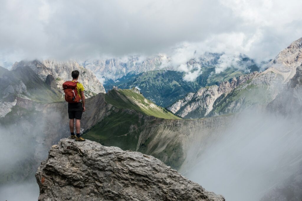 A hiker with a red backpack stands atop a rocky peak, gazing at misty mountains and lush valleys under a cloudy sky.