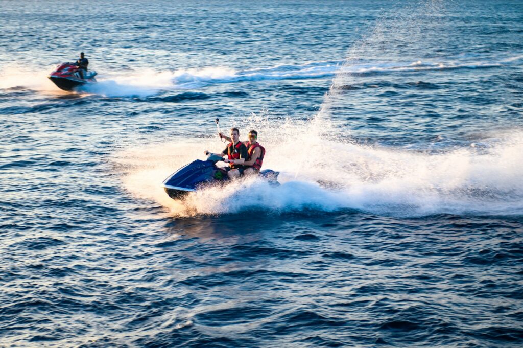 Two people joyfully ride a blue jet ski, creating splashes as they speed across the shimmering blue ocean under a clear sky.