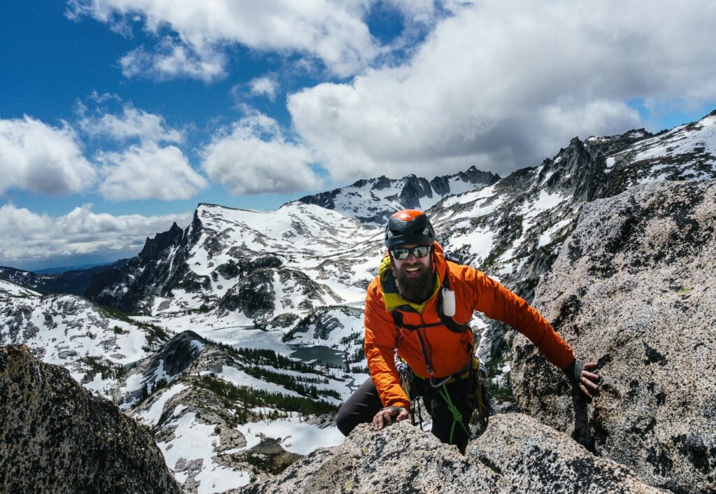 A climber in an orange jacket ascends rocky terrain surrounded by snow-capped mountains and a blue sky with scattered clouds.