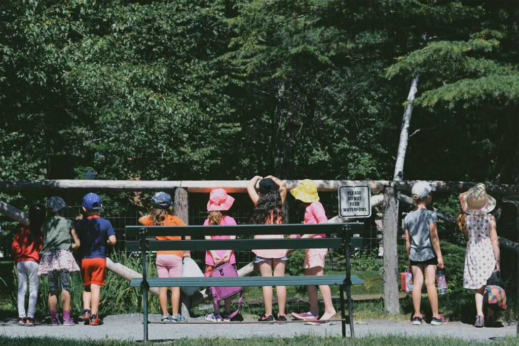 A group of children in colorful hats and outfits gathers by a fence, looking at a sign that says "Please do not feed waterfowl."