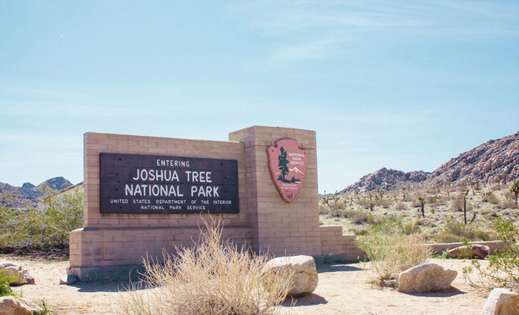 A sign welcoming visitors to Joshua Tree National Park, surrounded by desert landscape and rocky terrain under a clear blue sky.