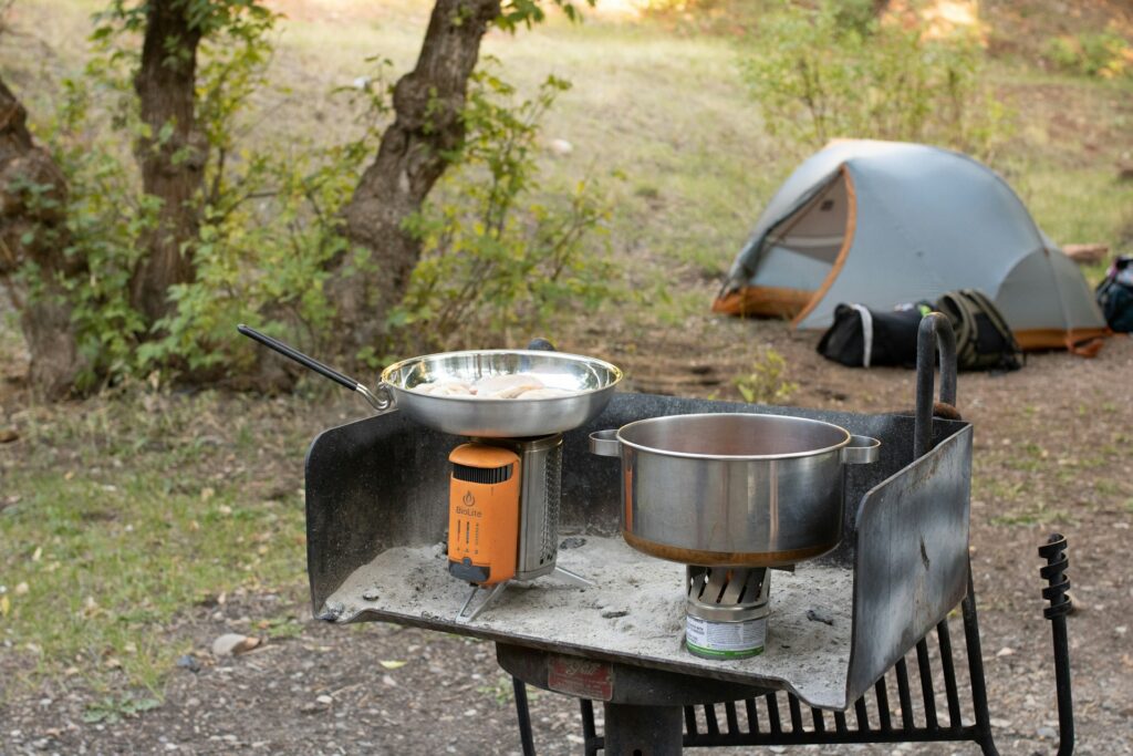 A portable camping stove with a skillet and pot cooking outdoors, with a tent visible in the background among trees.