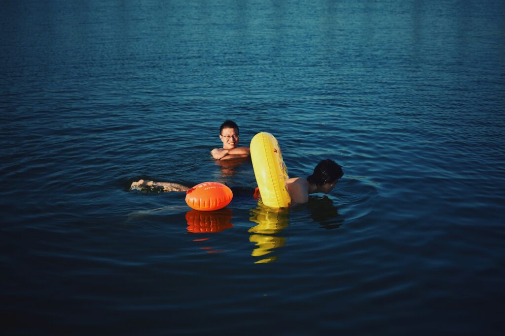 Two individuals swim in calm water, one using a yellow inflatable ring and the other a red float, under bright sunlight.