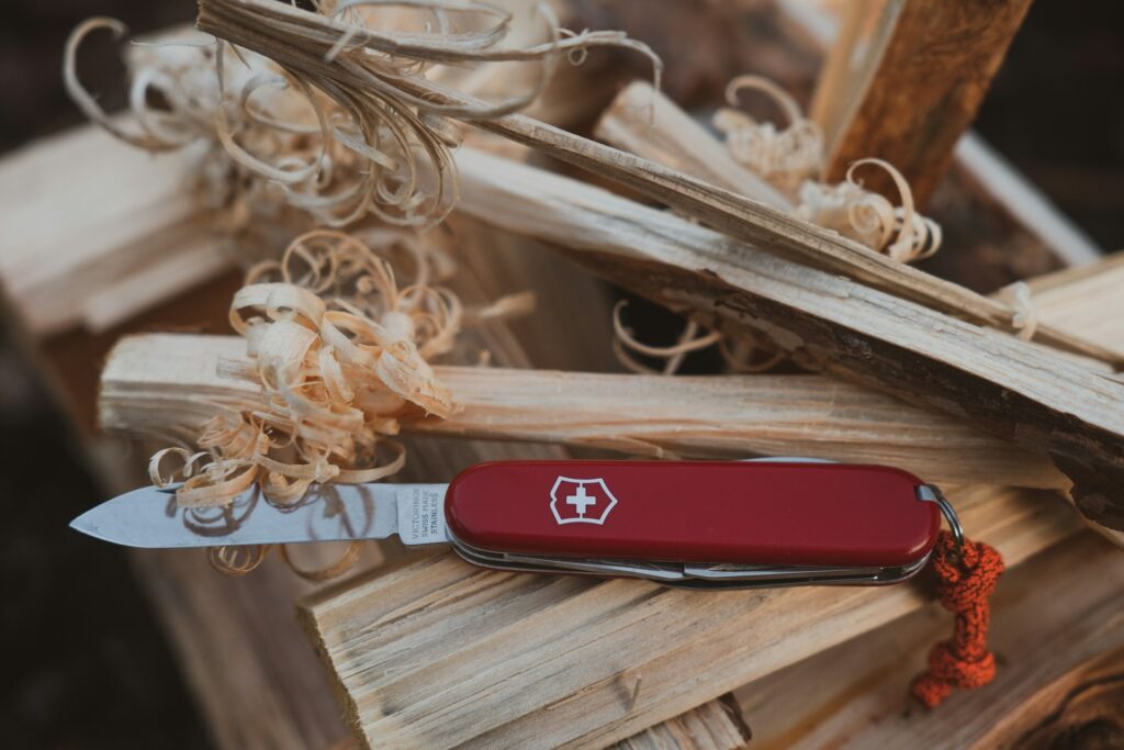 A red Swiss Army knife is resting on wooden logs, surrounded by wood shavings, showcasing its shiny blade and distinctive logo.