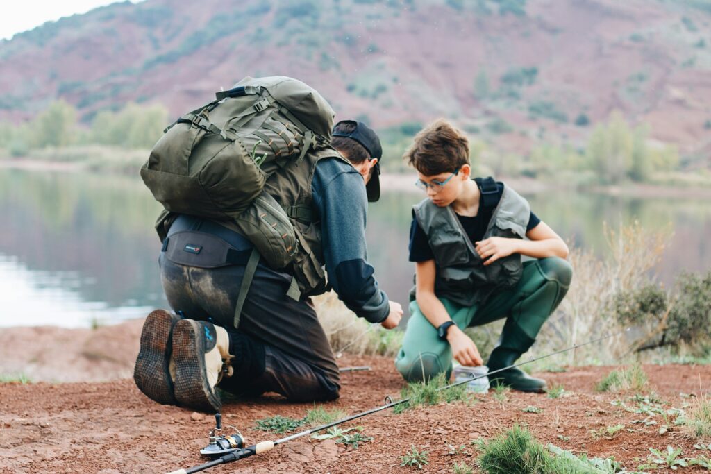 A person kneels beside a child near a lake, teaching them about fishing while surrounded by nature and camping gear.
